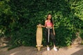 Stylish mulatto girl stands with longboard on wall background with ivy, looks away with serious face. Beautiful hispanic girl