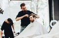 Stylish mustachioed barber dressed in a black shirt with a red bow tie scissors the hair of a young man in a barbershop