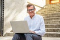 Happy attractive mature man working on laptop checking email sitting on stairs outdoors urban area Royalty Free Stock Photo