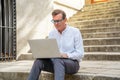 Stylish old man working on laptop surfing the internet sitting on stairs outdoors city in digital nomad Senior using modern Royalty Free Stock Photo