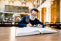 Stylish old senior elderly man with white beard and glasses working in an antique library with books, sitting at the Royalty Free Stock Photo
