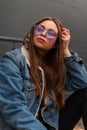 Stylish portrait cool young woman hipster in glasses in military hood in stylish blue vintage denim jacket near the gray building Royalty Free Stock Photo