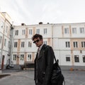 Stylish portrait of a young man with a hairstyle in fashion sunglasses in a cool leather black jacket with a trendy backpack on a Royalty Free Stock Photo