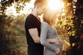 Stylish pregnant couple holding hands on belly in sunny light in autumn park. Happy young parents, mom and dad, hugging baby bump Royalty Free Stock Photo