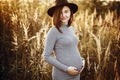 Stylish pregnant woman holding hands on belly and relaxing in sunny light in autumn park. Happy young parent, mom hugging baby Royalty Free Stock Photo