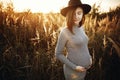 Stylish pregnant woman holding hands on belly and relaxing in sunny light in autumn park. Happy young parent, mom hugging baby Royalty Free Stock Photo