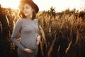 Stylish pregnant woman holding hands on belly and relaxing in sunny light in autumn park. Happy young parent, mom hugging baby Royalty Free Stock Photo