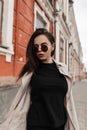 Stylish pretty urban young woman in black wear in fashionable spring youth trench coat in trendy sunglasses walk near vintage wall Royalty Free Stock Photo