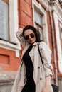Stylish pretty urban young woman in fashionable spring youth trench coat in trendy sunglasses stand near vintage wall in street. Royalty Free Stock Photo