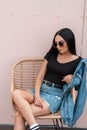 Stylish pretty young hipster woman in sunglasses in a black top in a vintage denim jacket is sitting on a straw chair in a cafe Royalty Free Stock Photo