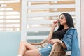 Stylish pretty young hipster woman in sunglasses in a black top in a vintage denim jacket is sitting on a straw chair in a cafe Royalty Free Stock Photo