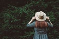 Stylish red-haired young woman in a straw hat and dress on a background of green trees. Shoot from the back. Place for text or Royalty Free Stock Photo