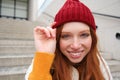 Stylish redhead girl in warm red hat, smiling relaxed, sitting with backpack on stairs near building, waits for someone Royalty Free Stock Photo