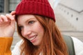 Stylish redhead girl in warm red hat, smiling relaxed, sitting with backpack on stairs near building, waits for someone Royalty Free Stock Photo