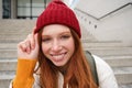 Stylish redhead girl in warm red hat, smiling relaxed, sitting with backpack on stairs near building, waits for someone Royalty Free Stock Photo