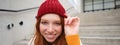 Stylish redhead girl in warm red hat, smiling relaxed, sitting with backpack on stairs near building, waits for someone Royalty Free Stock Photo