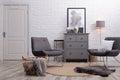 Stylish room interior with grey chest of drawers and chairs near white brick wall Royalty Free Stock Photo