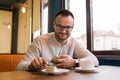 Stylish satisfied attractive businessman from Europe on a break eats sweet cheesecake food and drinks coffee sitting at a table in