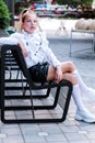 A stylish schoolgirl in a white blouse, leather shorts, socks and sports shoes sits in the park on an iron bench