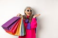 Stylish senior asian woman in sunglasses going shopping on holiday sale, holding paper bags and smiling, standing over Royalty Free Stock Photo