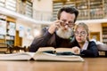 Stylish senior man grandfather reads interesting book to his pretty granddaughter, sitting together at the table in old