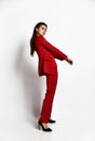 Sexy brunette woman is posing sideways to us in stylish red office pantsuit jacket, trousers and high-heeled shoes