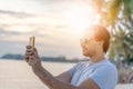 Stylish sexy young brunette man with a beard in sunglasses on a tropical beach with a mobile phone in his hands at sunset Royalty Free Stock Photo