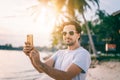 Stylish sexy young brunette man with a beard in sunglasses on a tropical beach with a mobile phone in his hands at sunset Royalty Free Stock Photo