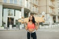 Stylish skater girl in trendy street clothes stands with longboard in hands on background of street and building and looks into