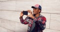 Stylish smiling african man taking selfie picture by phone in baseball cap, plaid shirt on city street over gray wall Royalty Free Stock Photo