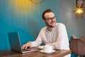 Stylish smiling fellow European businessman in a white shirt and glasses with a beard sits at a table in a cafe and drinks coffee