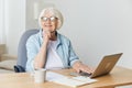 a stylish, successful elderly woman with gray hair works from home on a laptop and looks at the camera with a pleasant Royalty Free Stock Photo