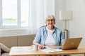 a stylish, successful elderly woman with gray hair works from home on a laptop and looks at the camera with a pleasant Royalty Free Stock Photo