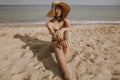 Stylish tan girl in hat sitting on beach. Fashionable young woman in straw hat, relaxing on sandy beach near sea in sunny light. Royalty Free Stock Photo