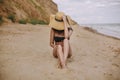 Stylish tanned girl in hat sitting on beach. Fashionable young woman covering with straw hat, relaxing on sandy beach near sea. Royalty Free Stock Photo