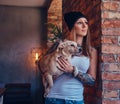 A stylish tattoed blonde female in t-shirt and jeans holds a cute dog. Royalty Free Stock Photo