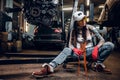 Stylish tattooed girl holding a big wrench sitting on a hydraulic hoist with a suspended car engine in the workshop Royalty Free Stock Photo