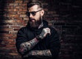 A stylish tattooed guy in a black hoodie and sunglasses. Studio photo against brick wall