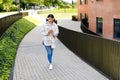 Stylish teen lady in wireless headphones texting on smartphone while walking in the urban city area or college campus Royalty Free Stock Photo