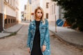 Stylish urban pretty young woman in a black stylish dress in a fashionable long denim jacket posing in a city on the street