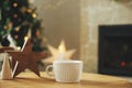 Stylish white cup with wooden star decor on table against stylish festive christmas tree with golden lights and cozy fireplace. Royalty Free Stock Photo