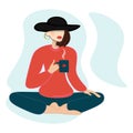 Stylish woman in hat drinking coffee and sitting in lotus position. Yoga practice, relaxation.