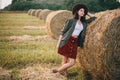 Stylish woman in hat standing at hay bale in summer evening in field. Tranquility. Rural slow life