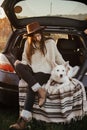 Stylish woman in hat and sweater sitting and hugging cute dog in car trunk in evening autumn field. Road trip with pet.Young Royalty Free Stock Photo