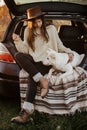 Stylish woman in hat and sweater sitting and hugging cute dog in car trunk in evening autumn field. Road trip with pet.Young Royalty Free Stock Photo