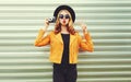 Stylish woman with retro camera taking picture wearing yellow jacket, black round hat on metal wall Royalty Free Stock Photo