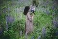 Stylish woman in rustic dress walking among lupine meadow, atmospheric image. Young female in linen dress gathering flowers in