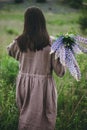 Stylish woman in rustic dress walking among lupine meadow, atmospheric image. Young female in linen dress gathering flowers in