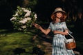 A stylish woman in a straw hat and a straw bag poses with a bouquet of white lilac in a Sunny spring Park. A quiet portrait of a Royalty Free Stock Photo