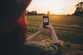 Stylish woman taking photo of sunset on phone in summer field. Atmospheric beautiful moment Royalty Free Stock Photo